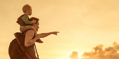 Moški, ki nosi majhnega otroka na ramenih, kaže proti obzorju ob sončnem zahodu.